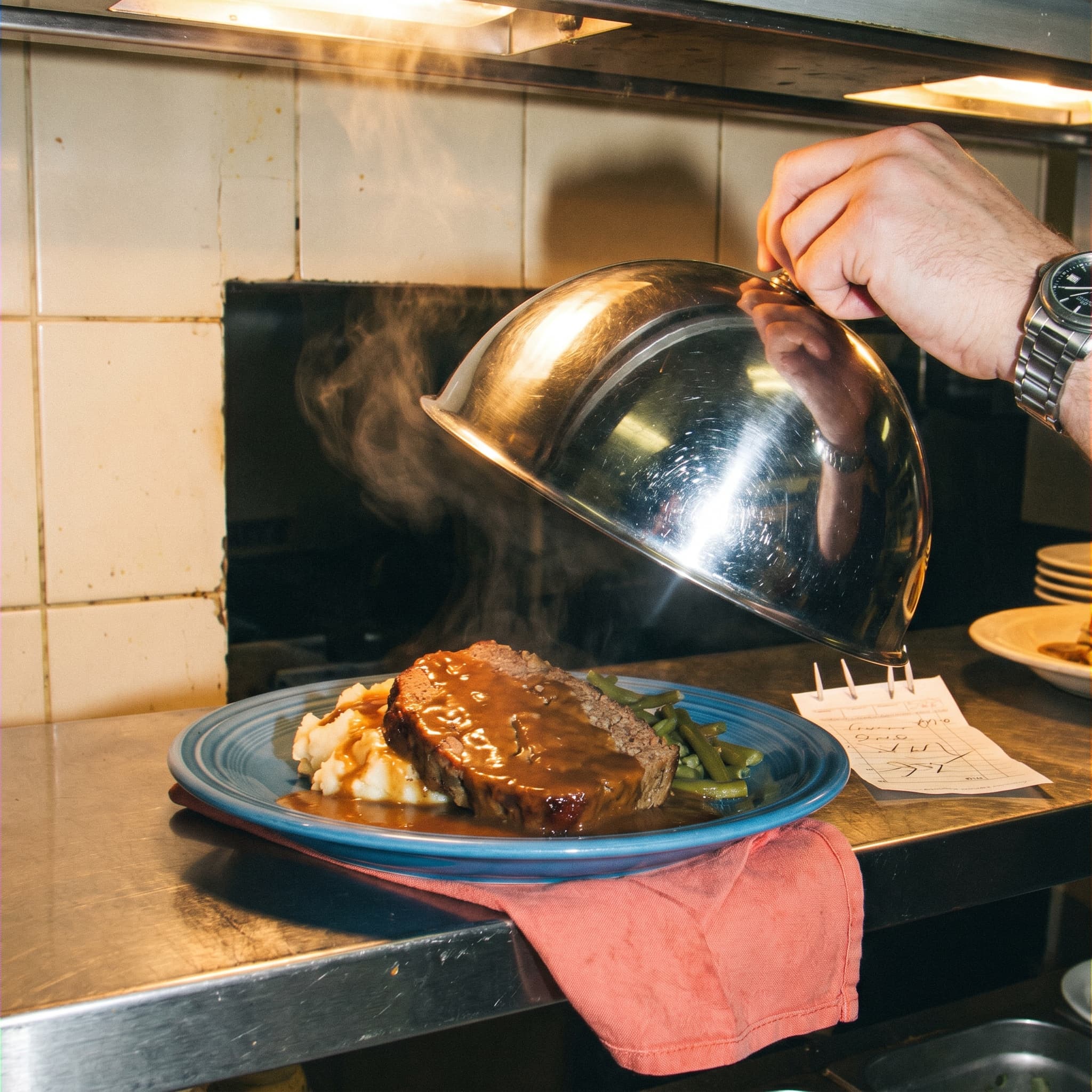 A classic blue-plate diner special under a chrome cloche, captured with hard flash on a counter.